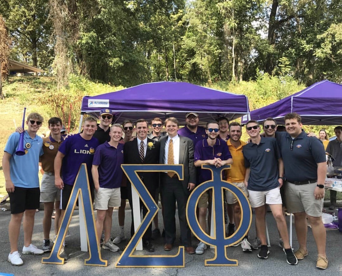 Members of Lambda Sigma Phi gather under a pop-up tent at a tailgate and pose behind large wooden Lambda Sigma Phi letters